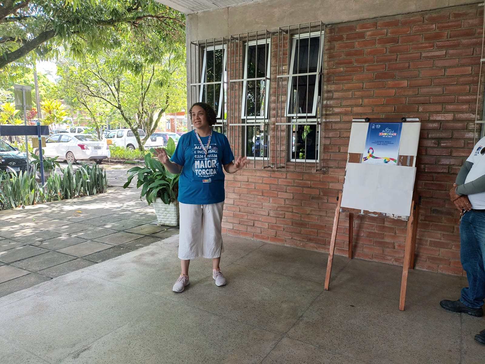 Mulher vestida com camisa azul gesticula ao lado do cartaz da UAEADTec em apoio ao Dia Mundial de Conscientização sobre o Autismo na área externa de um prédio